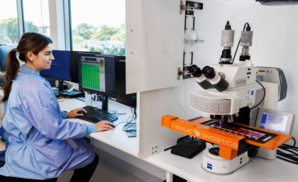 A female scientist in a blue lab coat sits at a computer beside a digital microscope scanning system. A scientist in SNP's Brisbane laboratory beside one of the digital pathology scanners.